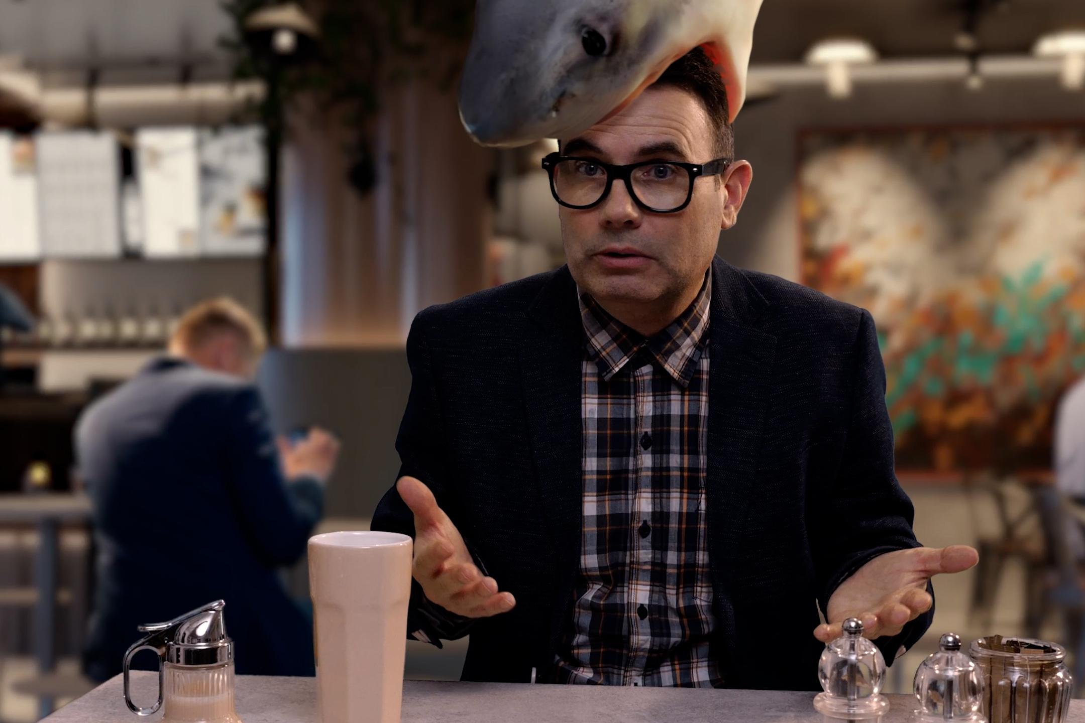 Eine Person sitzt an einem Tisch in einem modern eingerichteten Café oder Restaurant. Auf dem Kopf der Person befindet sich die realistisch dargestellte Figur eines großen Hais, der den Kopf von oben zu umschließen scheint. Vor der Person stehen ein helles Getränk in einem hohen Glas sowie mehrere Gewürzstreuer und eine kleine Karaffe. Im Hintergrund sind weitere Personen an Tischen zu sehen sowie eine Bar und dekorative Wandgestaltung.